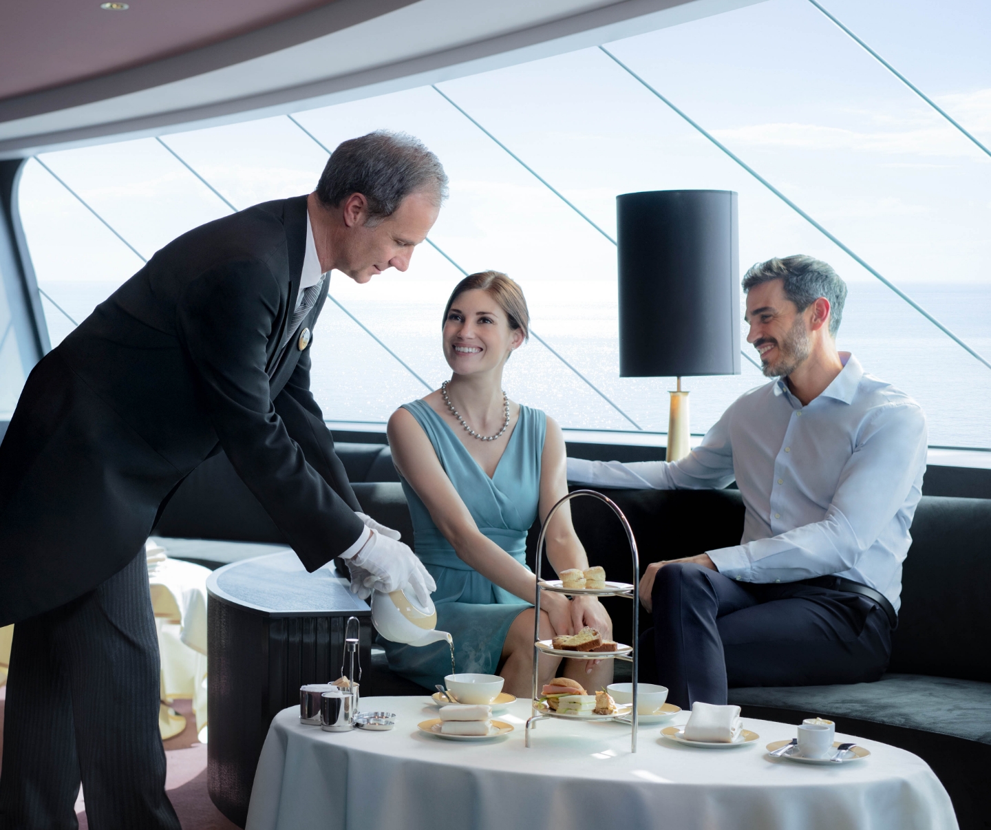 A MSC Yacht Club Butler in a distinguished black suit and white gloves serves tea with finger foods to a couple in a private yacht club area. They relax on a couch, savoring the moment while enjoying breathtaking ocean views. 