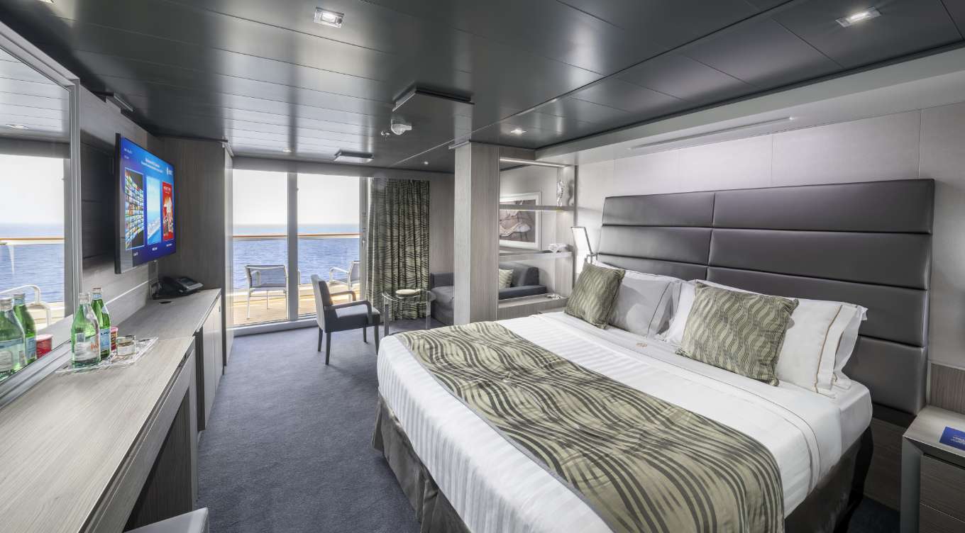 The Deluxe Suite on Seashore, is a well-lit and stylish indoor space in varying shades of grey exudes a modern and minimalist vibe. Notable features include a staircase hinting at spaciousness, a versatile table with chairs, and ample natural light through a window, creating an inviting atmosphere for your cruise vacation. 