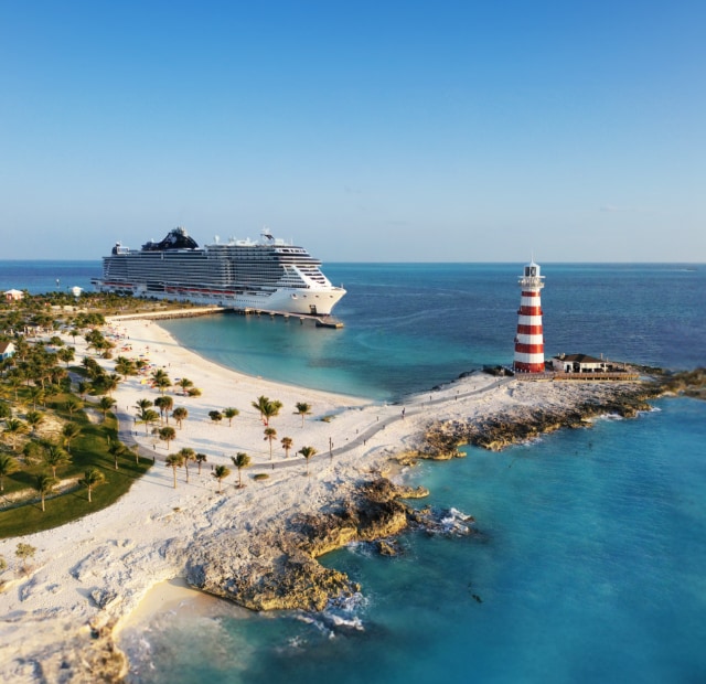 A beautiful shot of MSC Seascape docked at a Ocean Cays sandy beach, with a towering lighthouse in the background.