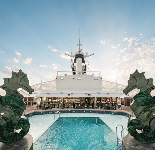 This is the private Yacht Club pool onboard MSC, featuring a spacious pool with easy-access stairs set against a vibrant blue sky with intriguing elements like a dinosaur figure, horses, and more, creating a unique and leisurely atmosphere. 