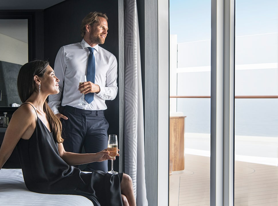 A man and woman each hold a glass of complementary champagne as they relax in their luxurious stateroom and enjoy their ocean cruise views through their balcony window. 