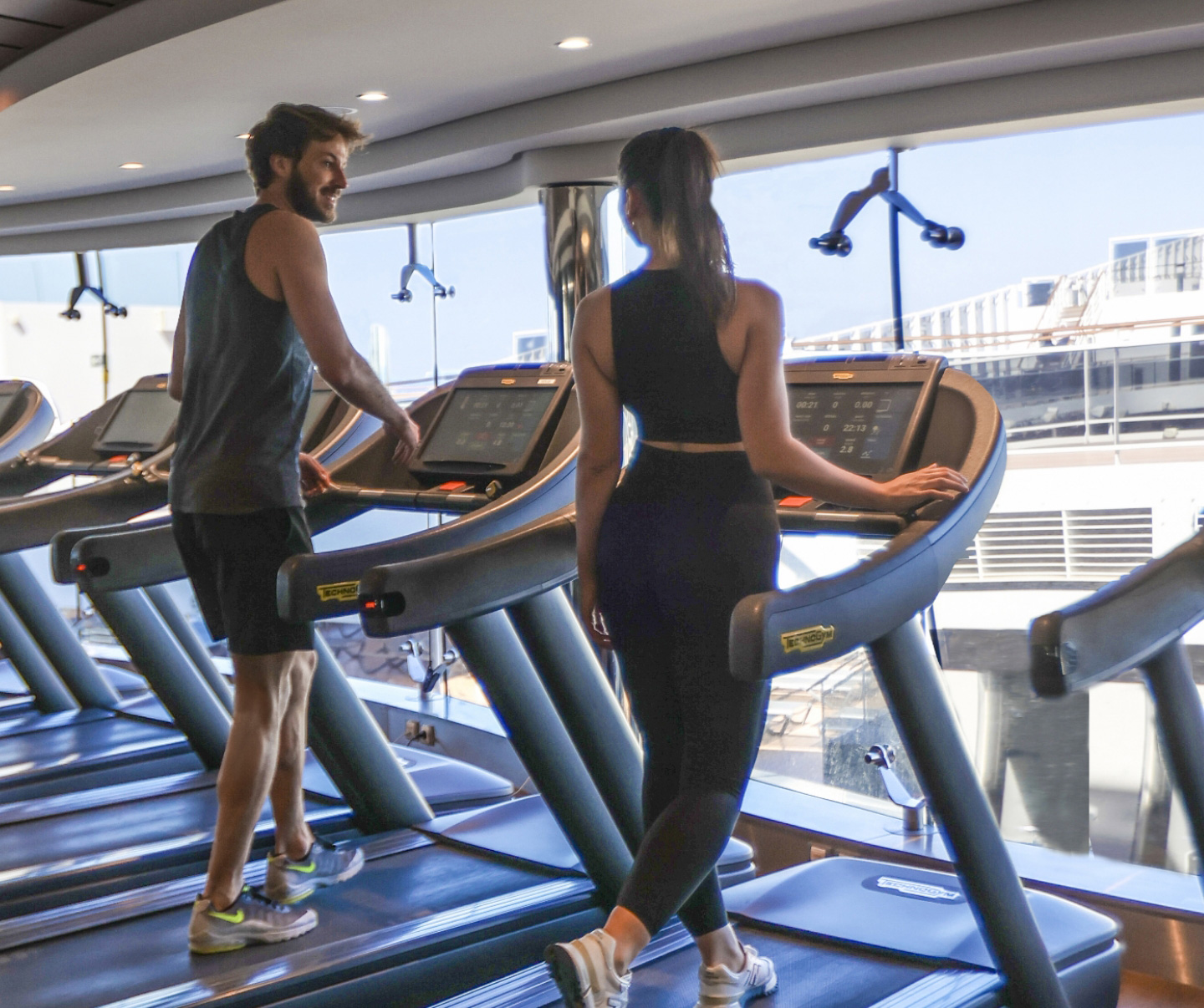 Enjoy a premium fitness experience on MSC Cruises as two individuals work out on treadmills in the well-appointed gym, offering scenic views of the ship and the expansive ocean beyond. 