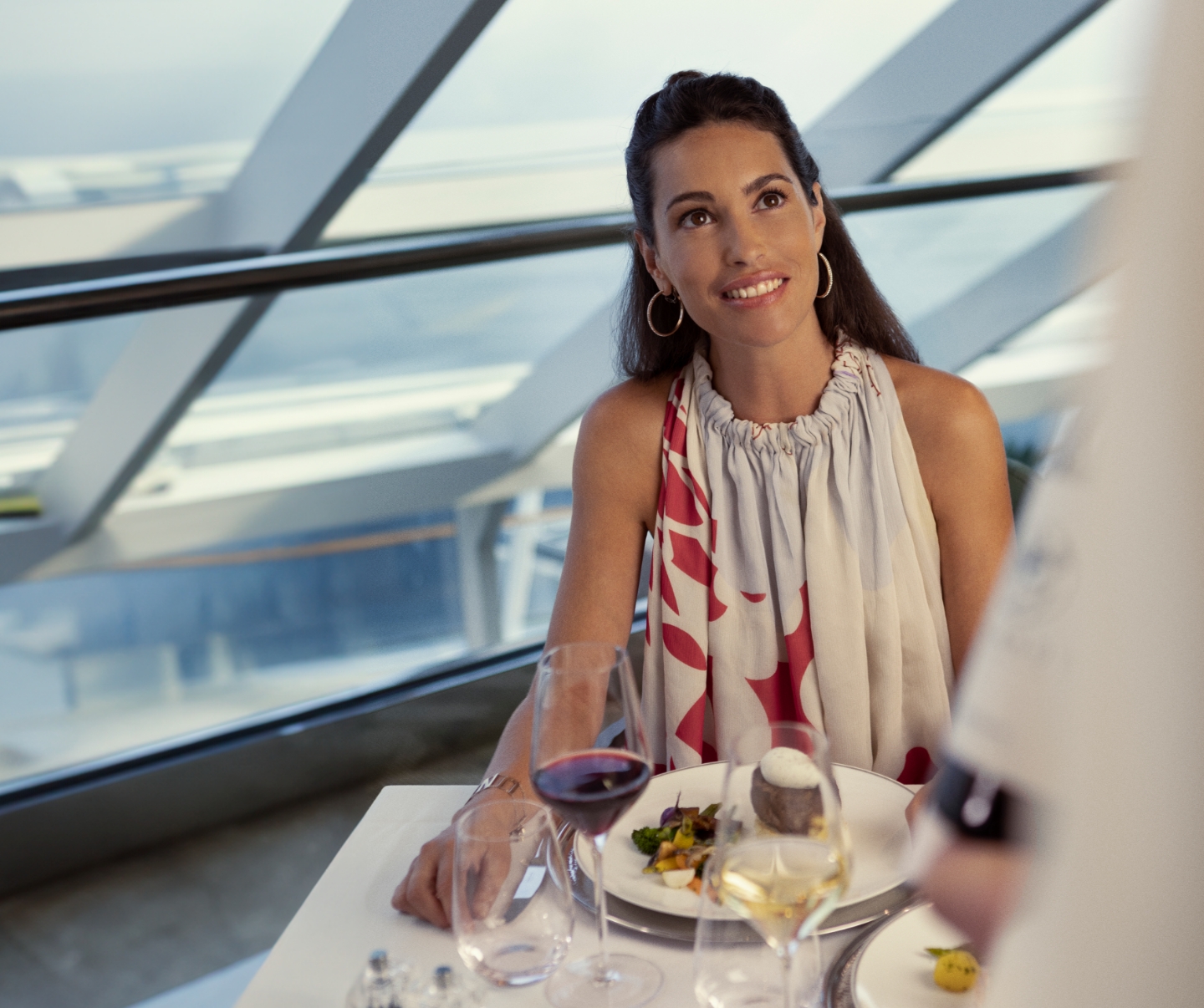 An elegant moment at the Yacht Club restaurant on your luxury cruise, as a delighted woman smiles while being served a gourmet dinner with a red and white wine pairing by the attentive Yacht Club staff, epitomizing the essence of dining and indulgence in luxury.